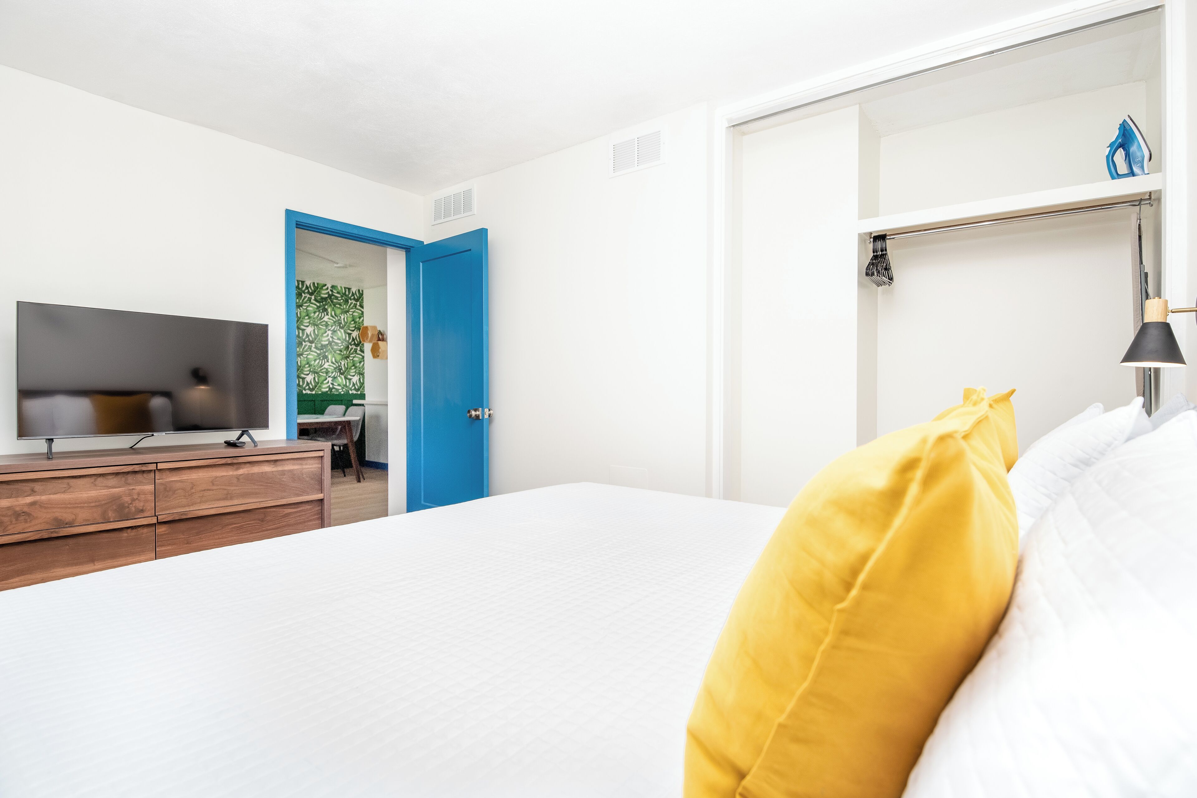 A modern bedroom with a bed, a TV on a wooden dresser, a blue door, and a shelf with a lamp.