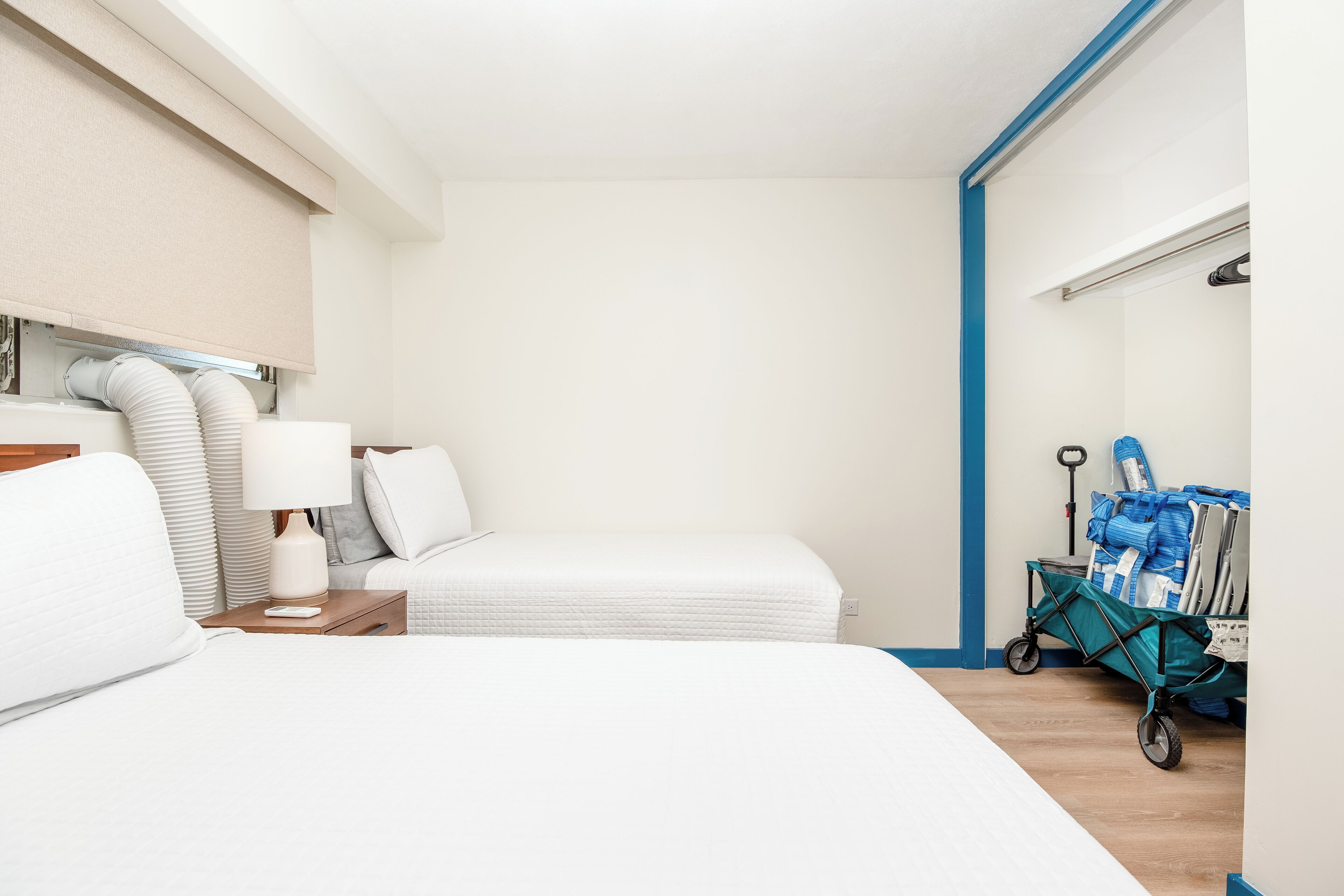 Quarto de hotel com cama, mesa de cabeceira, luminária, carrinho de mala e janela com persianas.