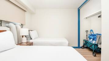 Quarto de hotel com cama, mesa de cabeceira, luminária, carrinho de mala e janela com persianas.