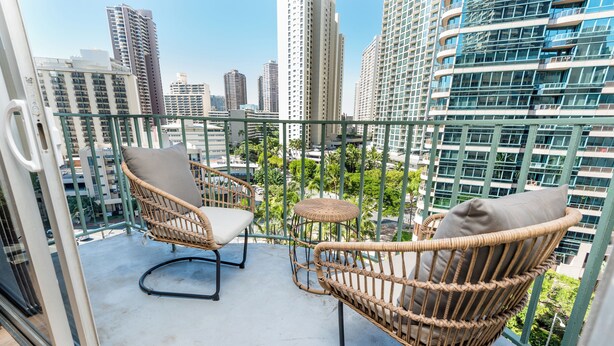 A balcony with wicker chairs and a small table, overlooking a cityscape with tall buildings. - Royal Aloha by Tropical Escapes Realty (Honolulu)