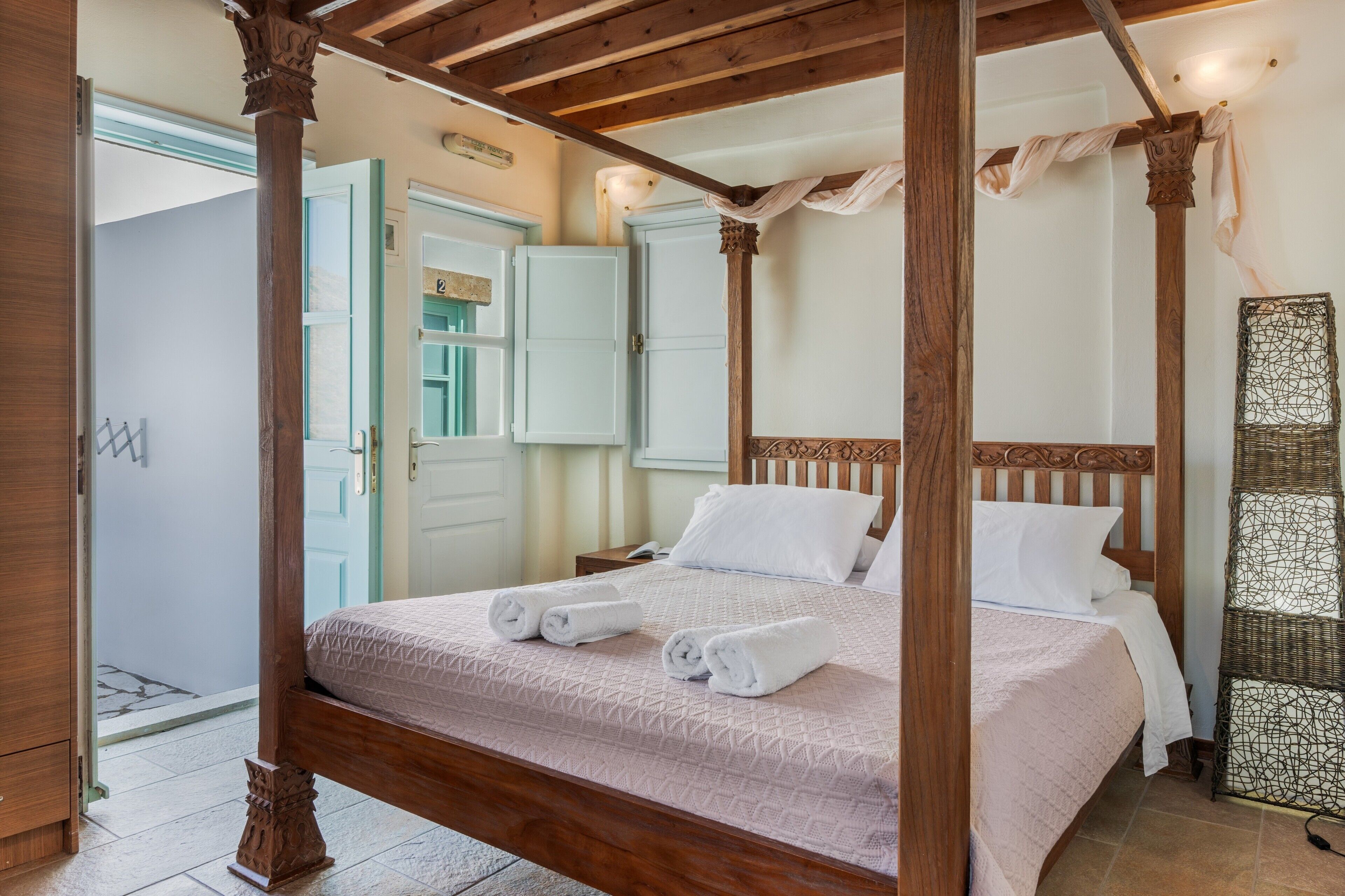 Un lit à baldaquin avec du linge de lit blanc, une tête de lit en bois et une table d’appoint en osier.