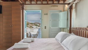 A bedroom with a bed, a wooden wardrobe, and a view of a town through an open door. - Room 'Double  Sea View No 1' with Sea View, Wi-Fi and Air Conditioning (Patmos)
