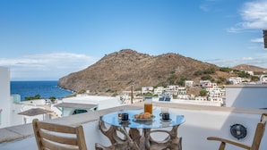 A table with a glass top and wooden chairs, a pitcher, and two cups on a balcony overlooking a coastal town with white buildings and a hill in the background. - Room 'Double  Sea View No 1' with Sea View, Wi-Fi and Air Conditioning (Patmos)