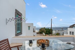 Un balcon avec une table en verre, deux chaises, un panier en osier rempli de viennoiseries et une vue sur la mer.