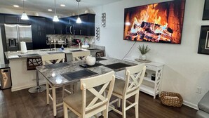 Una cocina moderna con armarios oscuros, encimera de mármol e iluminación colgante. Un televisor de pantalla plana grande muestra una escena de fuego.