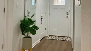 Un pasillo con paredes blancas, suelo de madera, una planta en maceta, un espejo y una puerta con paneles de vidrio esmerilado.