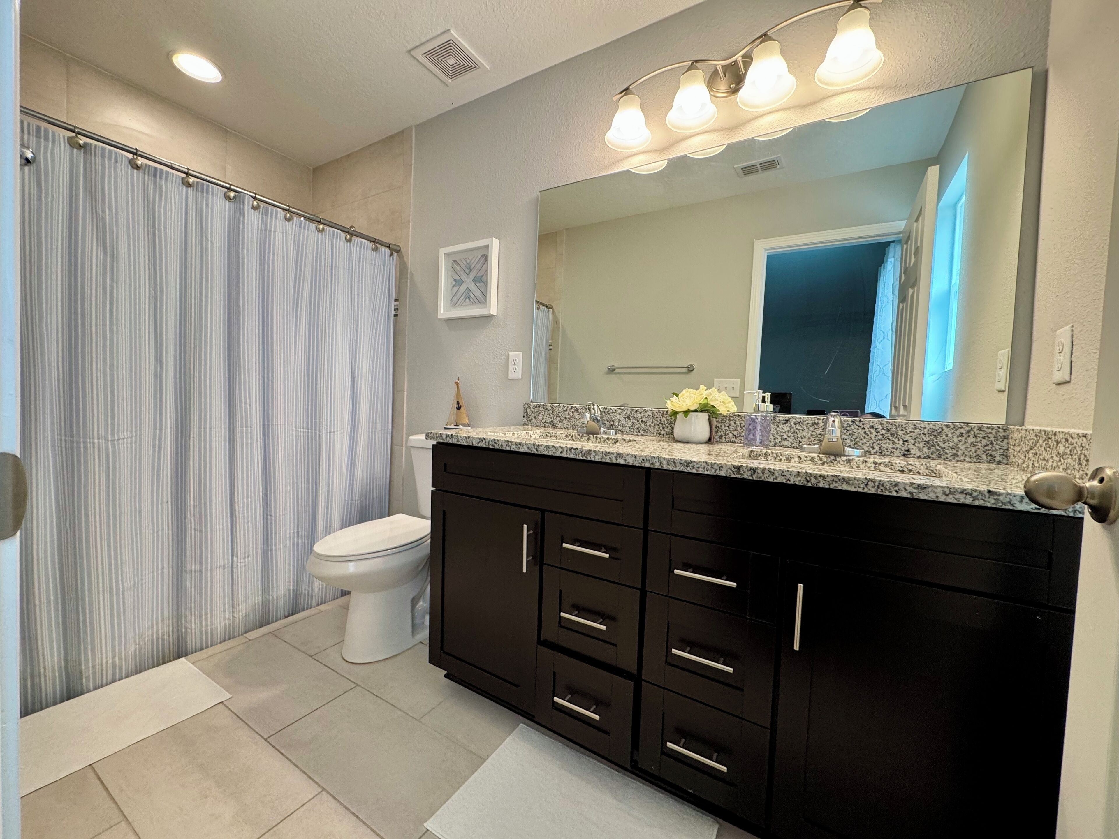 A bathroom with a shower, toilet, vanity with dark cabinets, and a mirror with lights.