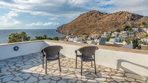A terrace with stone flooring and two wicker chairs, overlooking a coastal town with buildings and a hill.