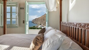 A bedroom with a bed, white curtains, and a view of the sea and hills.