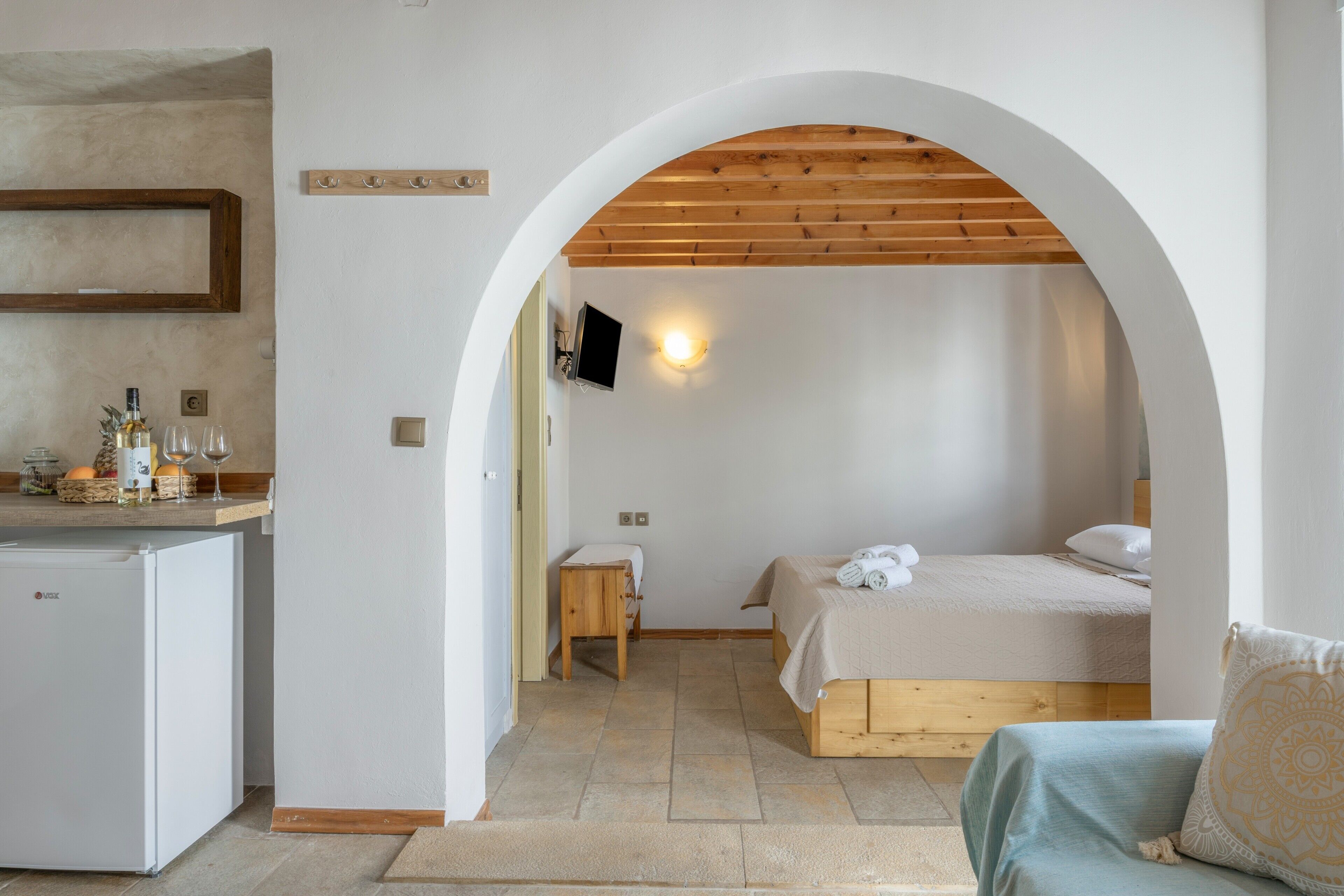 Une chambre comprenant un lit, une petite table de chevet, un téléviseur fixé au mur et un coin cuisine équipé d’un réfrigérateur.