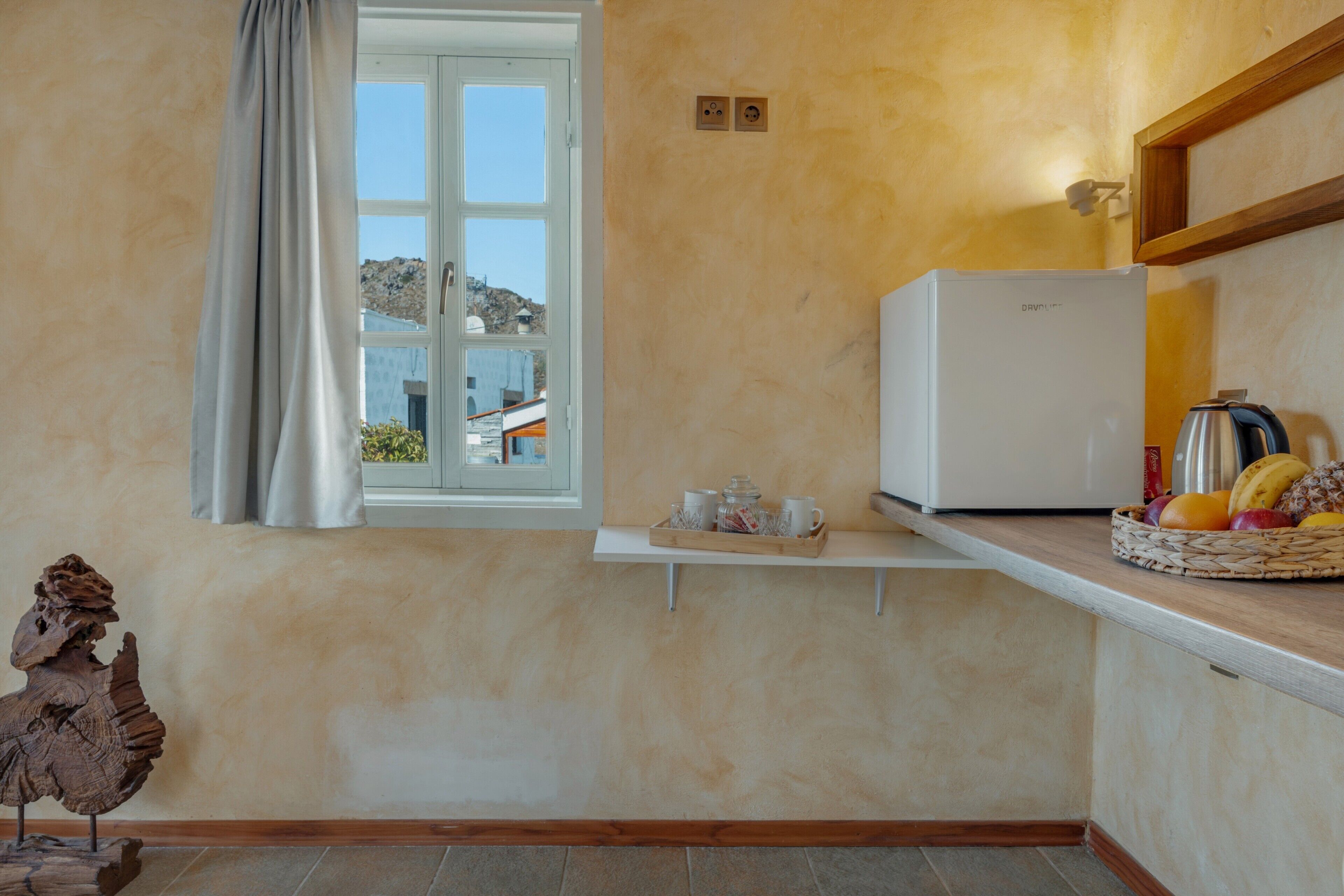 A kitchen with a refrigerator, a window with a view of buildings, a tray with cups and a basket of fruit.