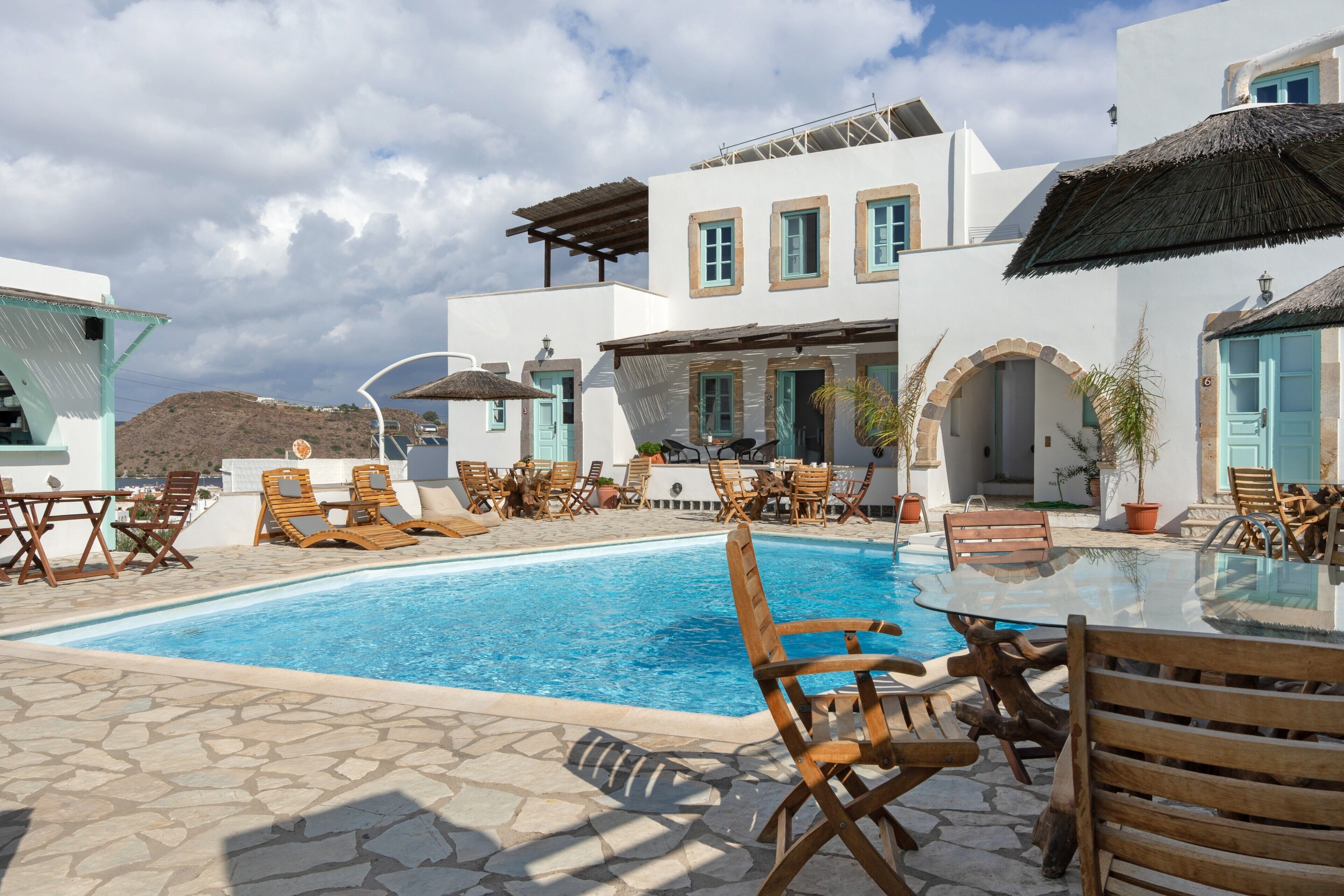A pool area with lounge chairs and a table, surrounded by white buildings with blue accents and a clear sky.