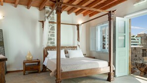 A four-poster bed with a stone wall and wooden beams, a nightstand with a vase, and a view of a stone building through an open door.