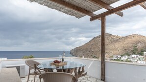 Eine Terrasse mit einem Glastisch, Korbstühlen und Blick auf das Meer und eine Hügel.