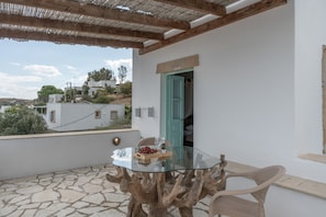 Une table à plateau en verre et à piétement en bois, une chaise en osier et une vue sur un bâtiment blanc avec une porte verte.