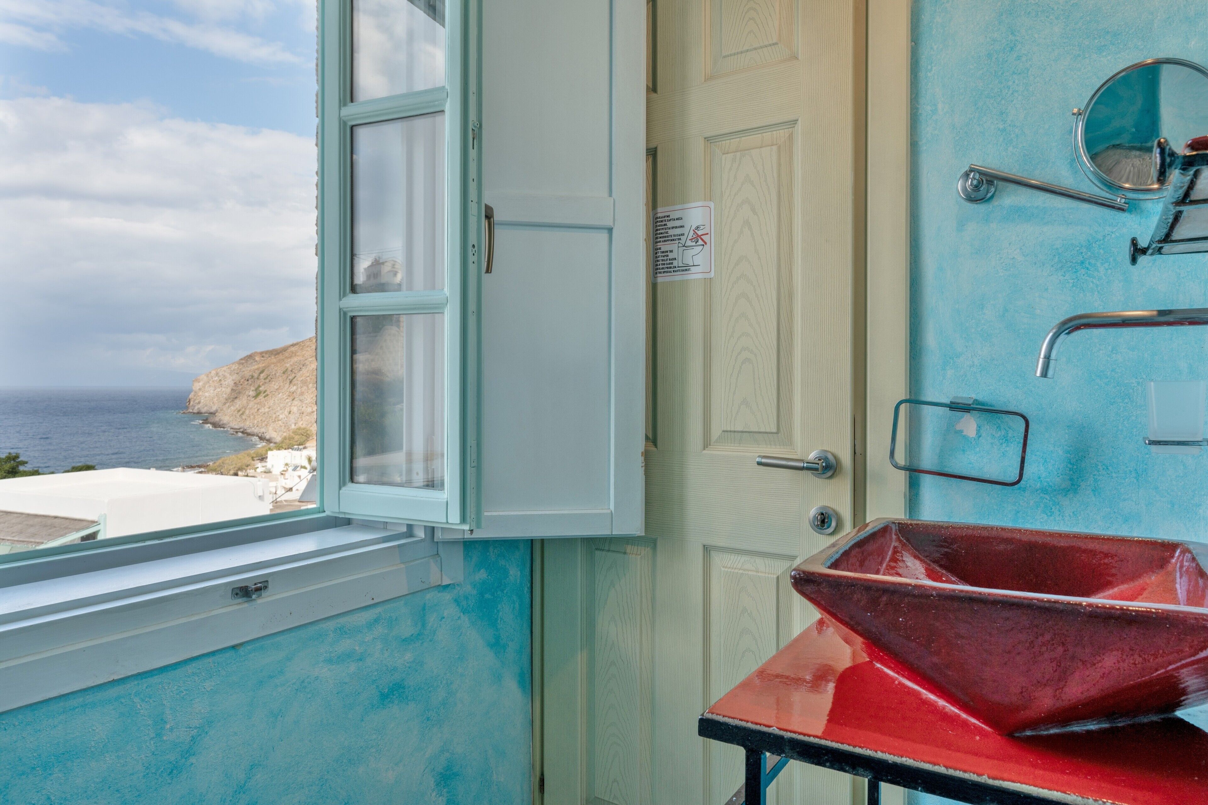 A bathroom with a view of the sea, a red basin, and a door with a sign.