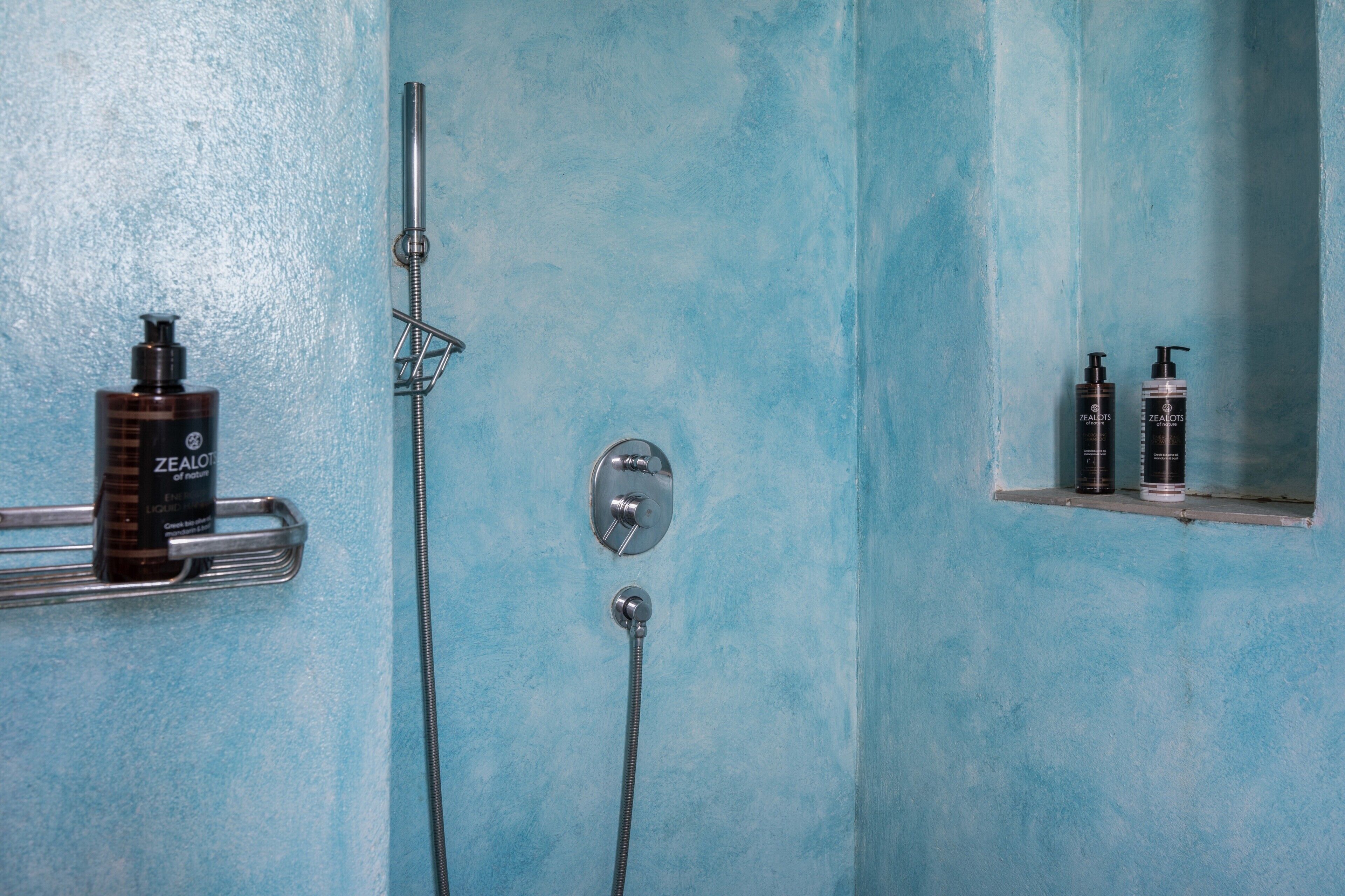 Een badkamer met een blauwe muur, een douche met een handdouche, en twee flessen Zealots body wash op een plankje.