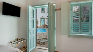Une chambre avec un lit, une télévision, une fenêtre avec des volets et une vue sur une piscine et des bâtiments à l’extérieur.