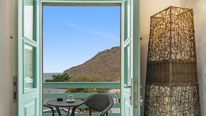 Une porte ouverte donnant sur un balcon où se trouvent un fauteuil en osier, une petite table et une vue sur une colline.