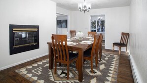 A dining room with a wooden table set for six, surrounded by six wooden chairs with dark cushions. A chandelier hangs from the ceiling, and a patterned rug covers the floor.