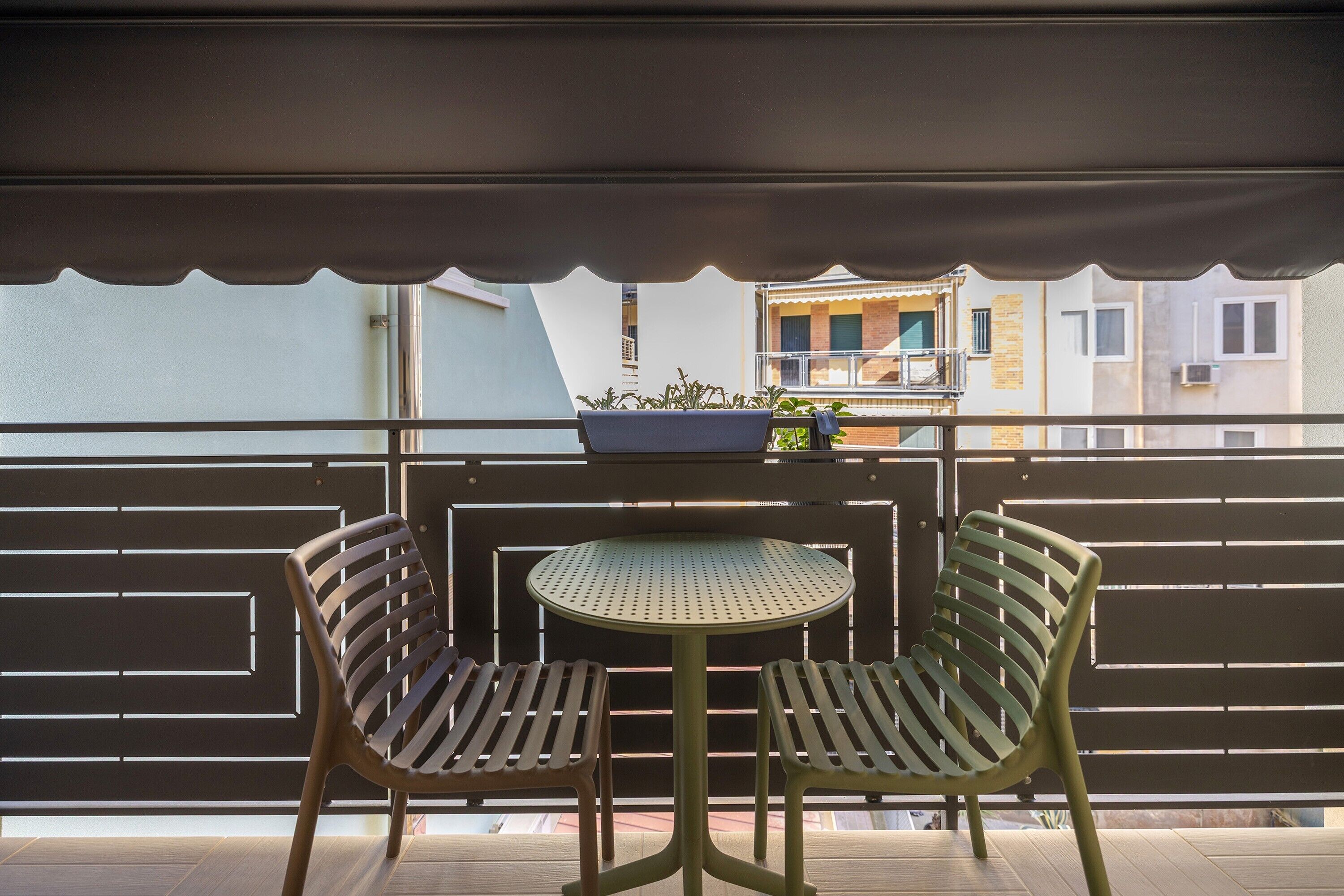 Ein Balkon mit zwei Stühlen und einem kleinen Tisch, Blick auf Gebäude, sowie ein überdachter Bereich mit Geländer.