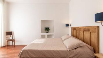 Un dormitorio con cama, una silla, una estantería con una planta y una lámpara de pared.