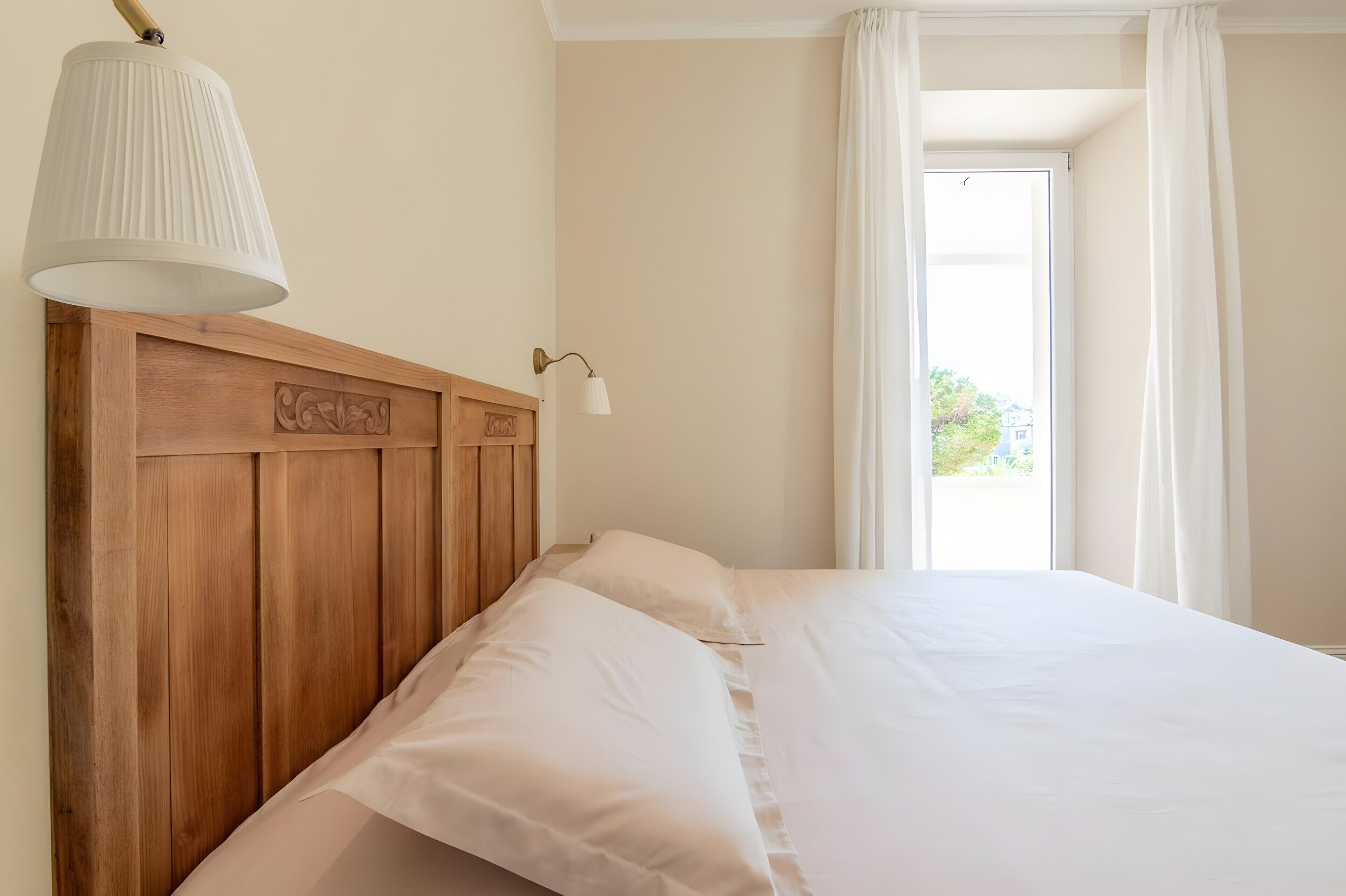 Ein Bett mit Holz-Kopfteil, einer weißen Lampe und einem Fenster mit Vorhängen.