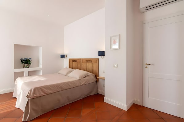 Une chambre à coucher comprenant un lit, une table de chevet, une lampe fixée au mur, une porte et une étagère sur laquelle est posée une plante.