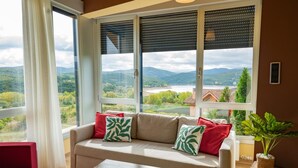 A living room with a sofa, red cushions, a wooden table, and a view of a lake and mountains through large windows. - Villa 'Casa El Rasillo' with Lake View, Private Terrace and Wi-Fi (El Rasillo de Cameros)