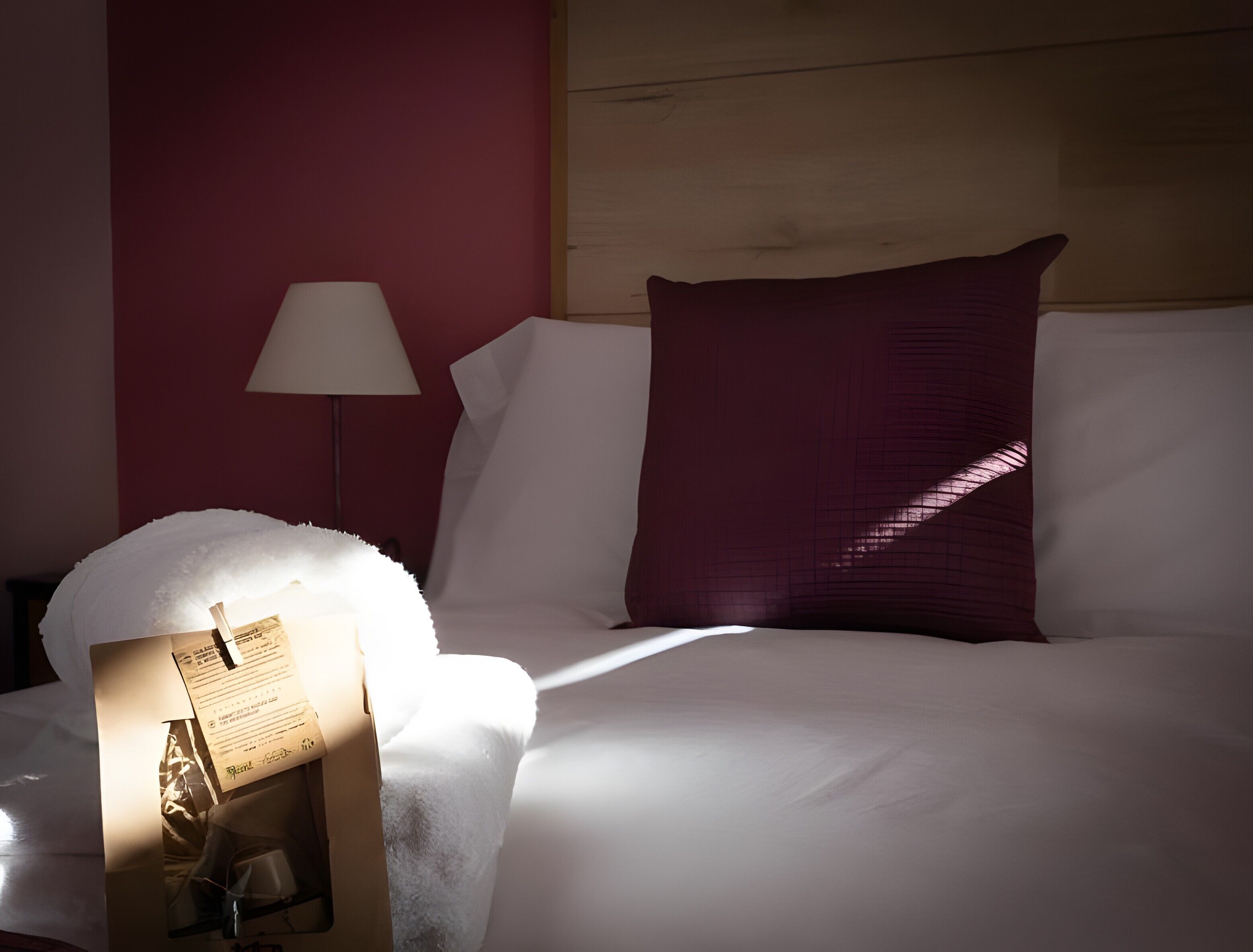 A neatly made bed with white bedding and a single red pillow, a bedside lamp, and a small table with a bottle and a glass.