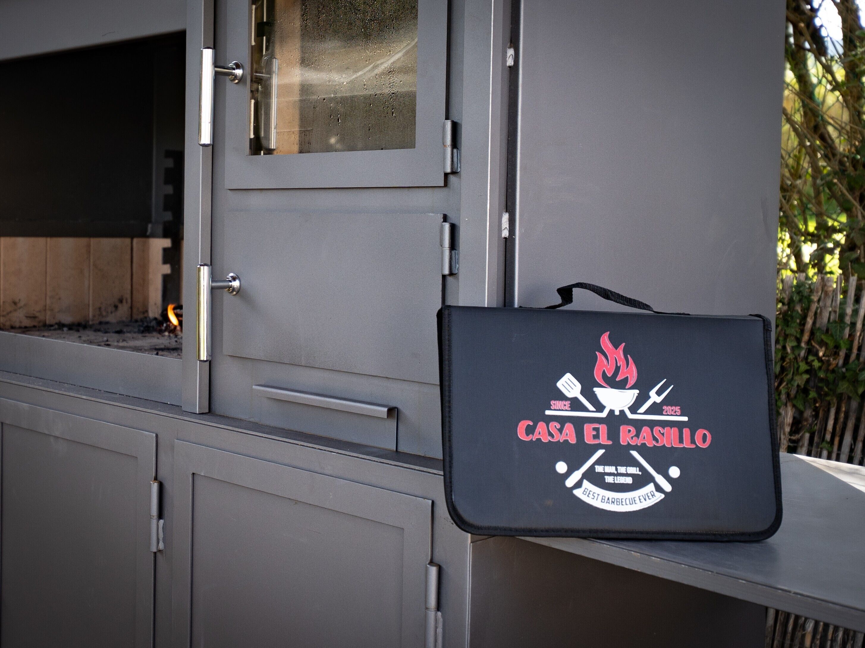 Una parrilla para barbacoa con un estuche negro identificado con 'Casa El Rasillo' y fuego visible en su interior.