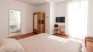Une chambre à coucher comprenant un lit, une armoire en bois, un bureau avec une chaise, un téléviseur et une salle de bain visible à travers une porte ouverte.