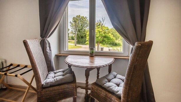 Deux fauteuils en osier avec des coussins gris, une petite table ronde en bois et une vue par une fenêtre ornée de rideaux.