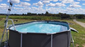 Une piscine extérieure avec un escalier, entourée d’une pelouse et d’un champ de maïs, sous un ciel partiellement nuageux.