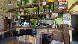 Un coin bar au style rustique, avec des étagères en bois, un tableau noir servant de menu et un mur en pierres en arrière-plan.