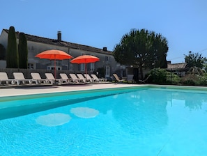 Una piscina con sillones de playa y sombrillas rojas, rodeada por un edificio con techo de tejas y un árbol.