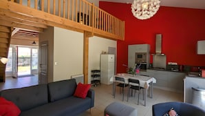 A modern living room with a grey sofa, a dining table, and a kitchen area with red walls and white cabinets. - Holiday Home 'La Bonne Chauffe' with Shared Pool, Private Terrace and Wi-Fi (Verrières)