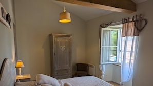 A bedroom with a bed, two bedside lamps, a wardrobe, a chair, and a window with curtains. - Holiday Home 'La Bonne Chauffe' with Shared Pool, Private Terrace and Wi-Fi (Verrières)
