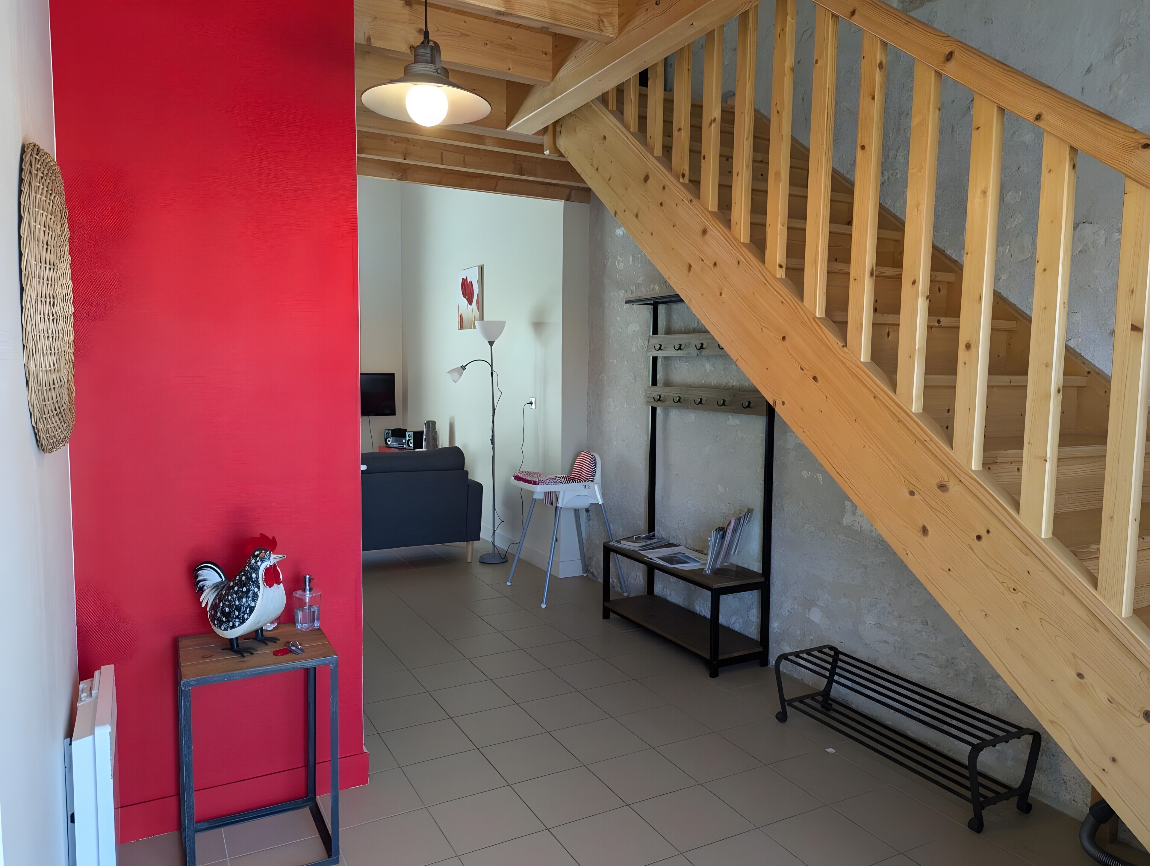 A living room with a wooden staircase, a red wall, a sofa, a table with a rooster decoration, and a floor lamp.