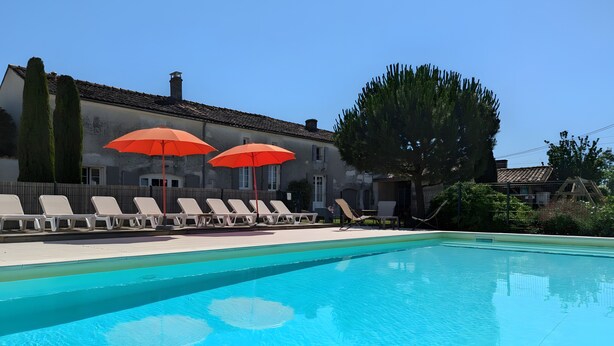A swimming pool with lounge chairs and red umbrellas, surrounded by a building with a tiled roof and a large tree. - Holiday Home 'La Bonne Chauffe - Serpentin' with Shared Pool, Private Terrace and Wi-Fi (Verrières)