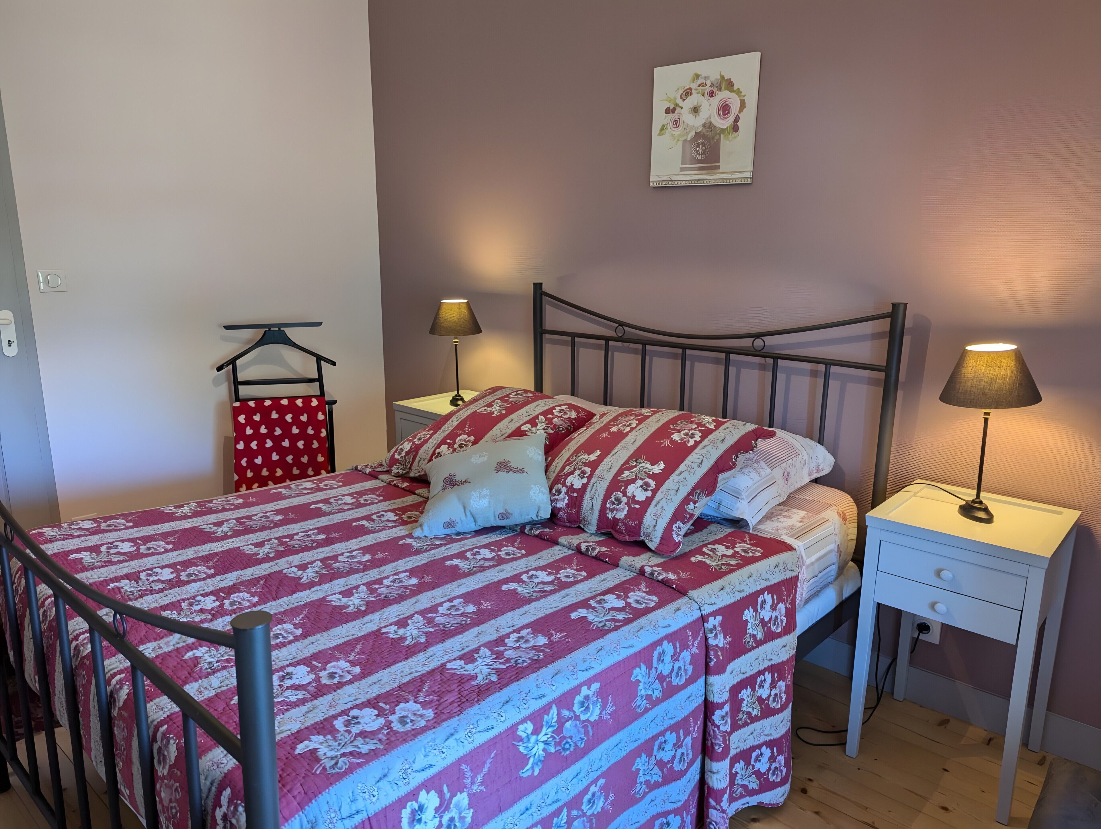 Un dormitorio con estructura de cama de metal, colcha roja y blanca con estampado floral, dos lámparas de noche, una mesita de noche blanca y un cuadro colgado en la pared.