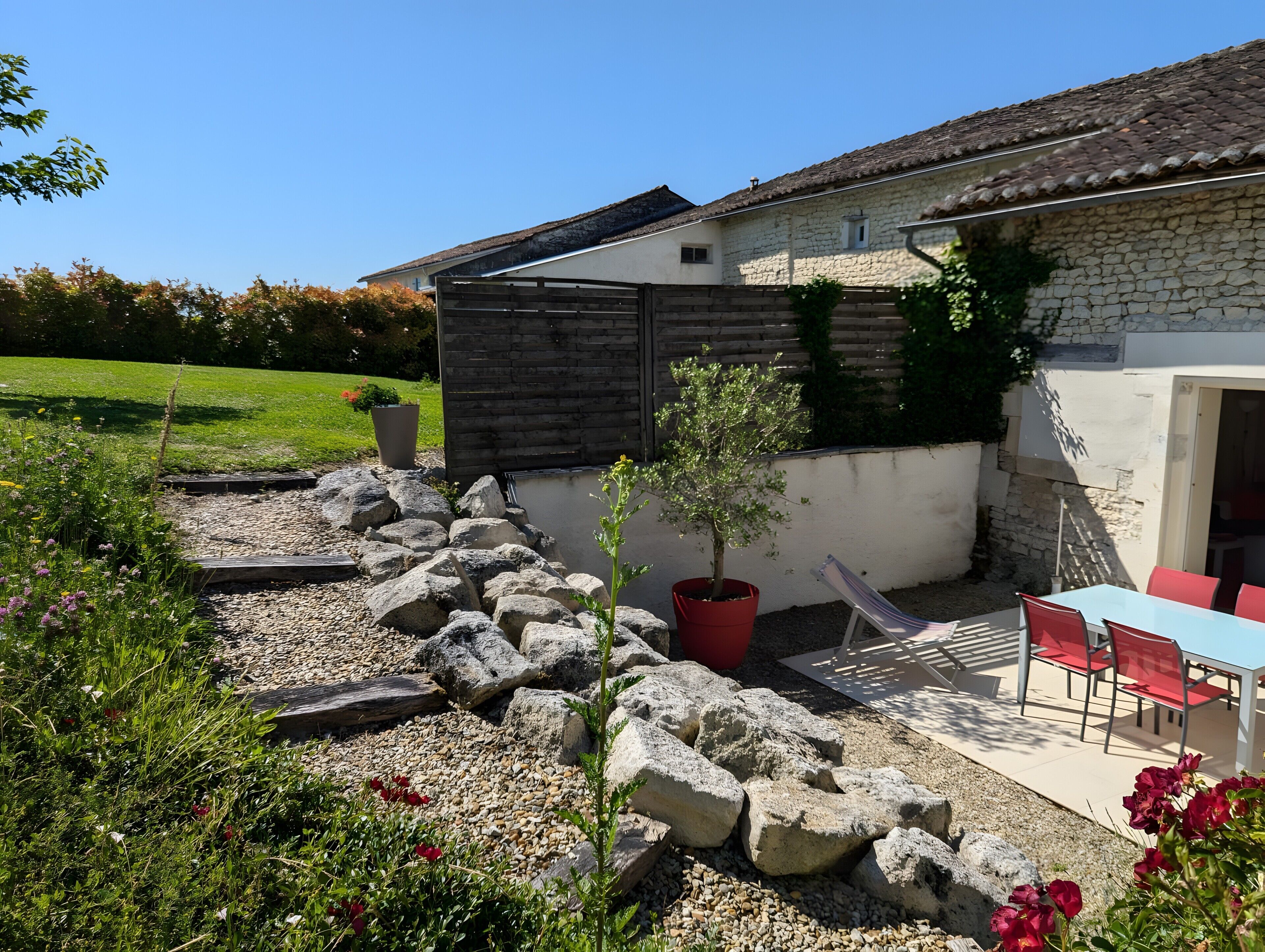 Un bâtiment en pierre avec un toit en tuiles, un jardin avec des allées de gravier et un espace détente avec des chaises rouges.