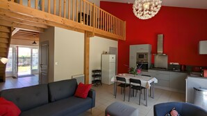 A modern living room with a grey sofa, a dining table, and a kitchen area with red walls and white cabinets. - Holiday Home 'La Bonne Chauffe-Col De Cygne' with Shared Pool, Private Terrace and Wi-Fi (Verrières)