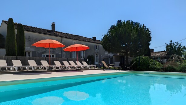 A swimming pool with lounge chairs and red umbrellas, surrounded by a building with a tiled roof and a tree. - Holiday Home 'La Bonne Chauffe-Col De Cygne' with Shared Pool, Private Terrace and Wi-Fi (Verrières)