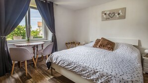 A bedroom with a bed, a small table, chairs, a window with curtains, and a wall decoration.