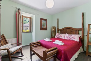 A bedroom with a wooden bed, a wooden chair, a table, a window with curtains, and a radiator.