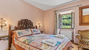 Une chambre avec un grand lit, une tête de lit en bois, une fenêtre donnant sur un paysage verdoyant et un fauteuil à motifs.