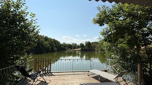 Une terrasse en bois, avec des chaises et une table en métal, donnant sur un lac paisible, le tout entouré d’arbres.