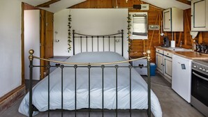 A bedroom with a metal bed frame, a wooden ceiling, a white wall, a small kitchen area with a sink and cabinets, and a window with curtains. - Chalet by Bramefort Pond (Champsac)