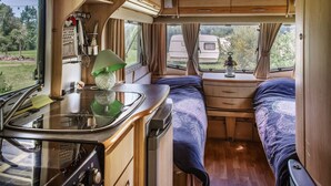 Ein kompaktes Wohnmobil-Interieur mit einer Küchenzeile, einem Bett mit gemustertem Bettbezug und einem Fenster mit Vorhängen.